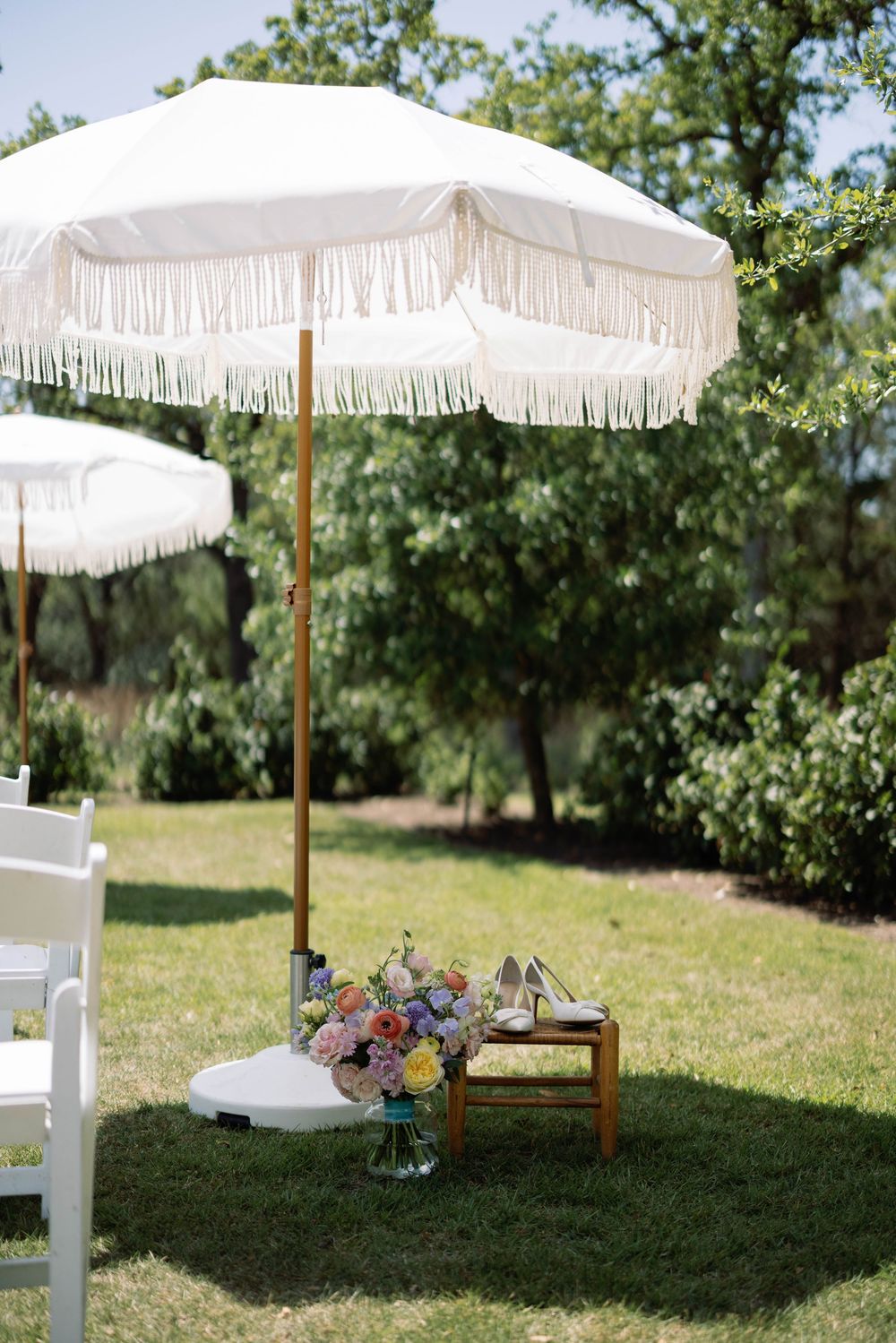 White fringed parasols line a grassy lawn at an outdoor wedding ceremony setup on a sunny summer day.