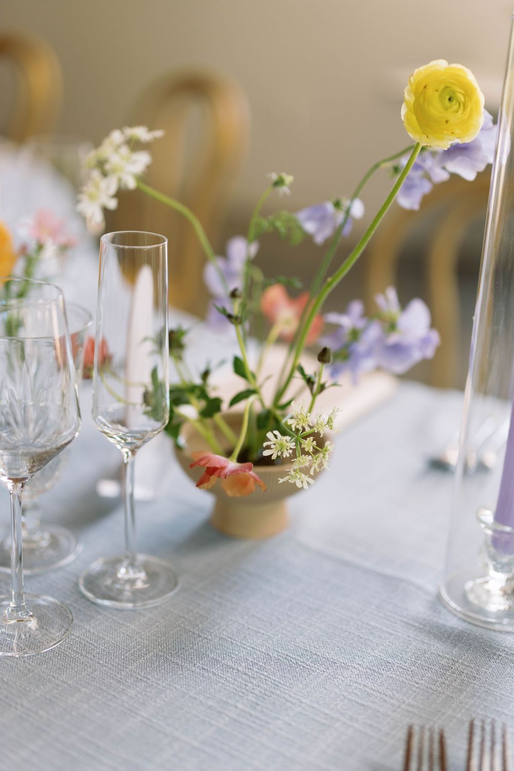 Delicate yellow ranunculus flowers and white blooms in glass vases on a gray linen tablecloth with crystal stemware.