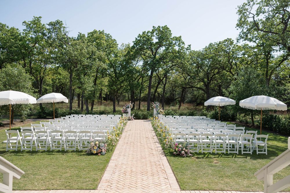 White wooden chairs arranged in rows for an outdoor wedding ceremony on a brick pathway surrounded by green trees.