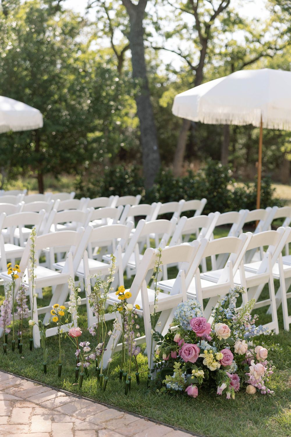 Garden wedding ceremony setup with white folding chairs adorned with fresh pink and yellow floral arrangements along the aisle.