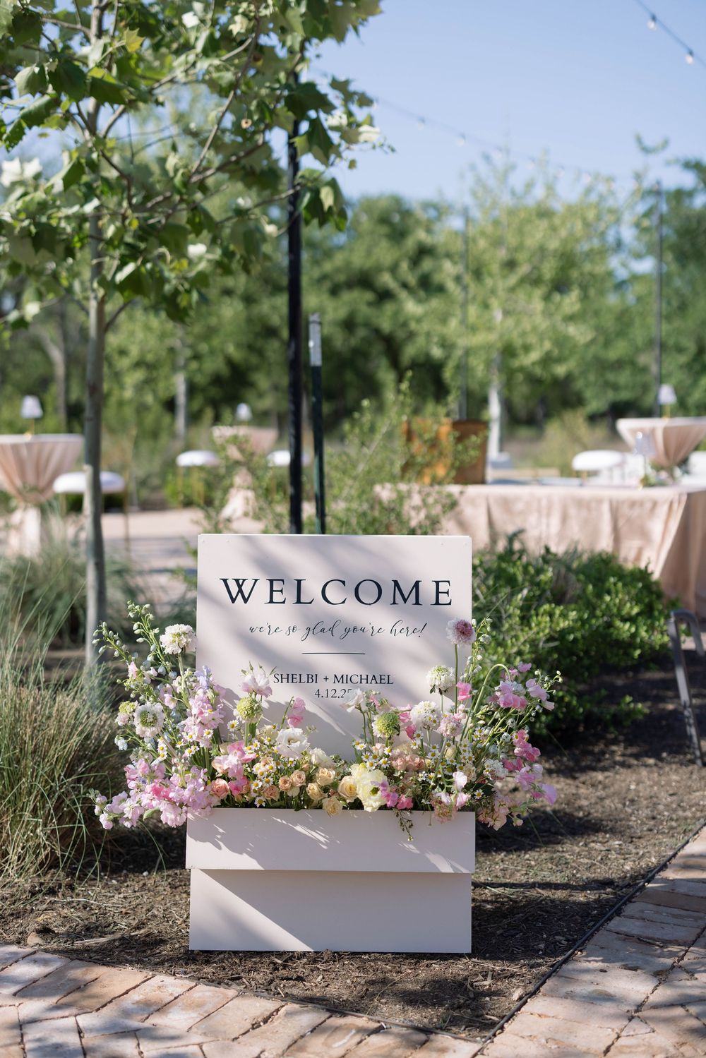A series of welcome signs surrounded by pink flowers and greenery in an outdoor garden setting.