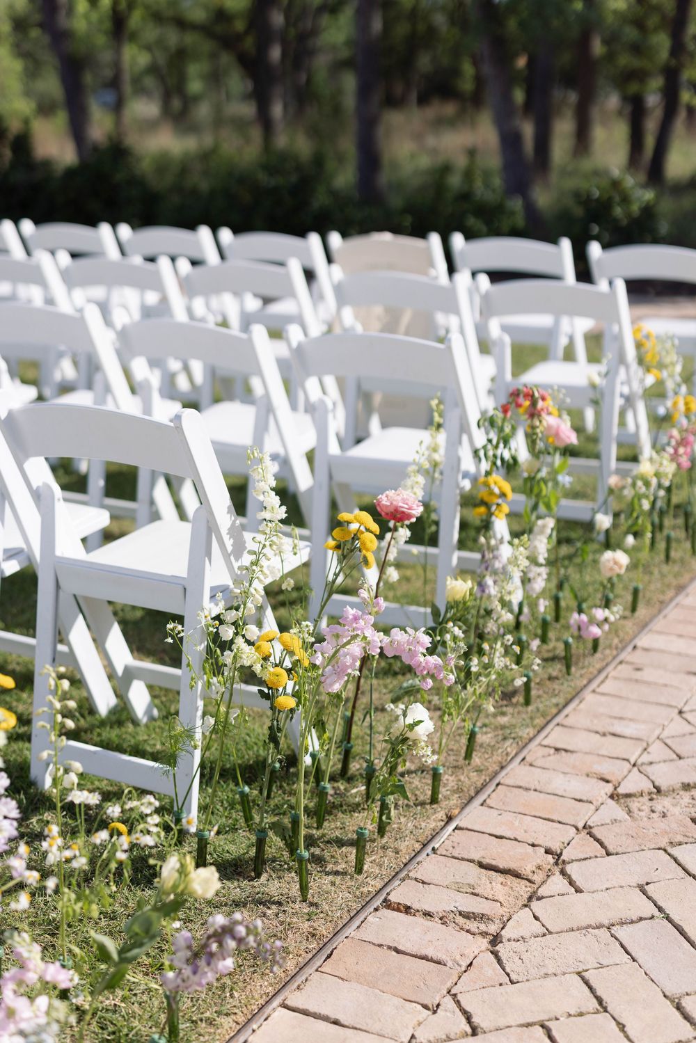 White folding chairs line a brick path with wildflowers creating a romantic outdoor wedding ceremony setup.