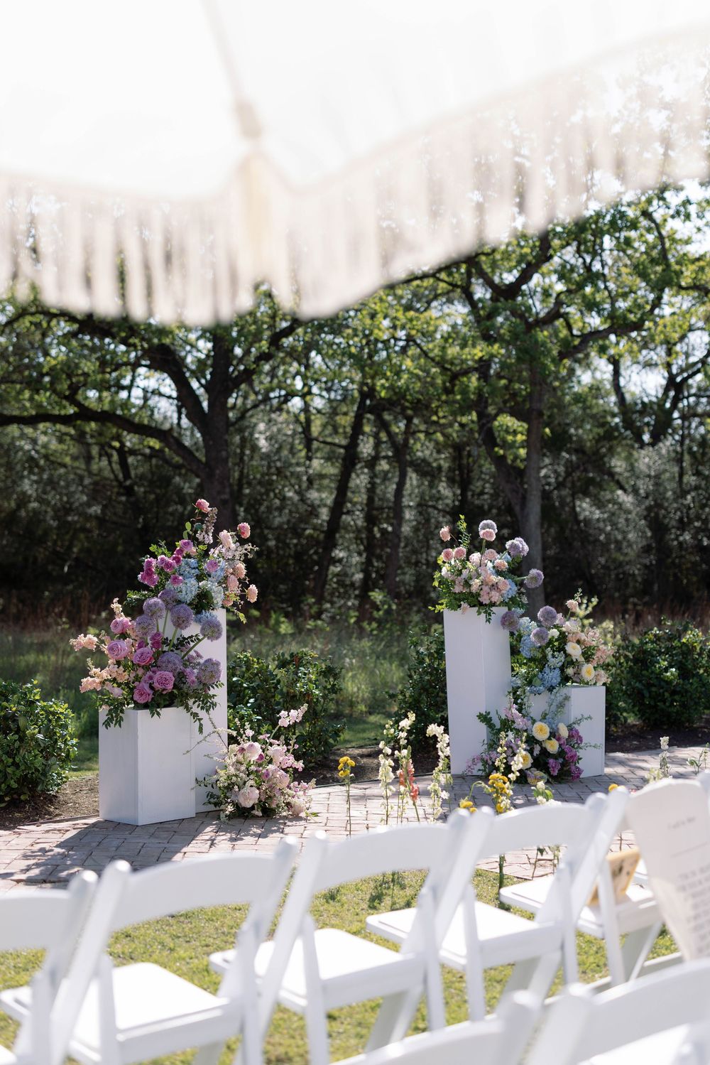 White folding chairs set up outdoors with tall floral arrangements on white pedestals for garden wedding ceremony.