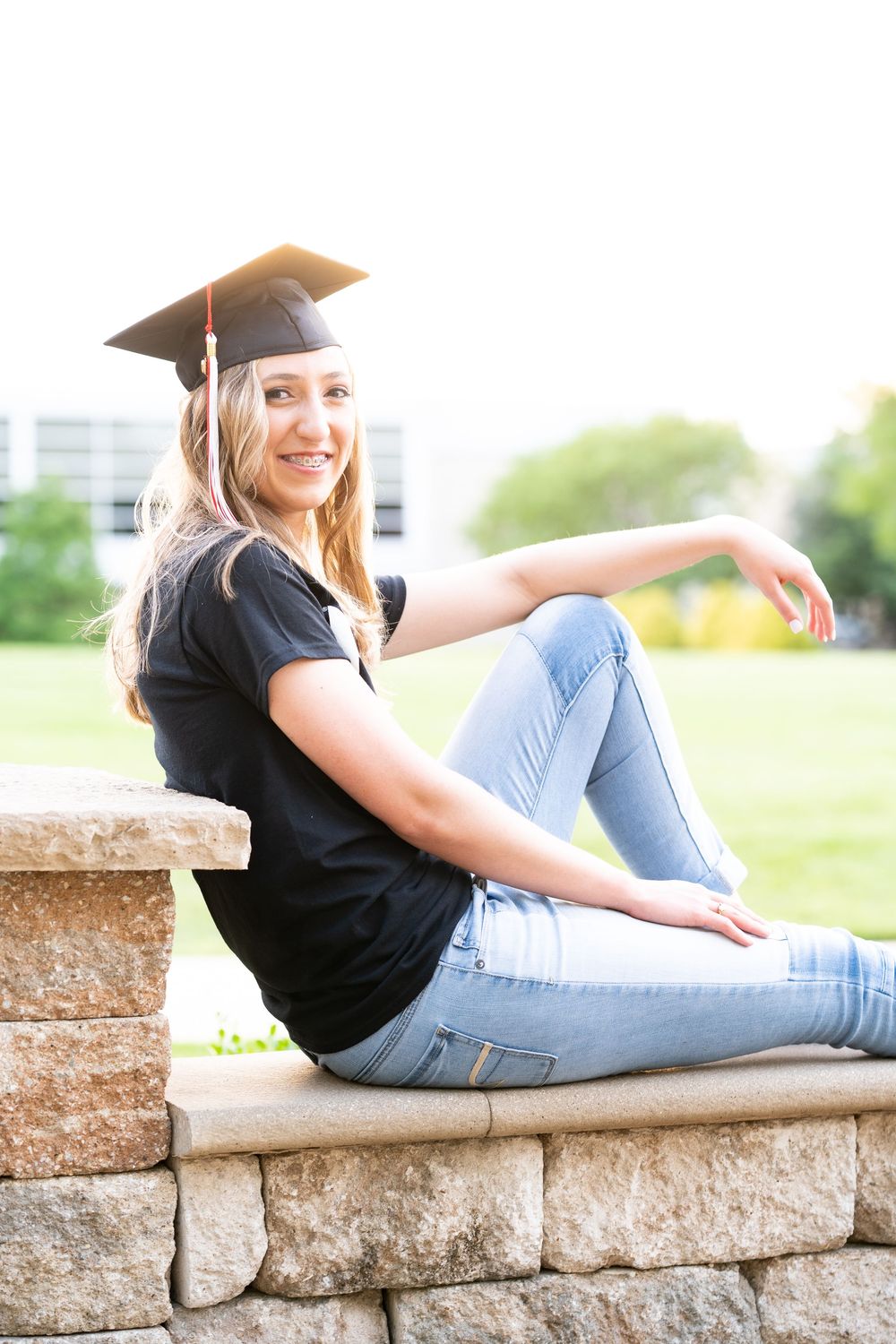 Graduation portrait of college student in cap sitting on a rock wall with arm over knee with green field in background.