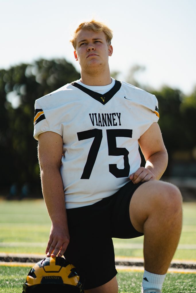 Senior portrait of high school football student wearing jersey kneeling on a football field with helmet at his feet.