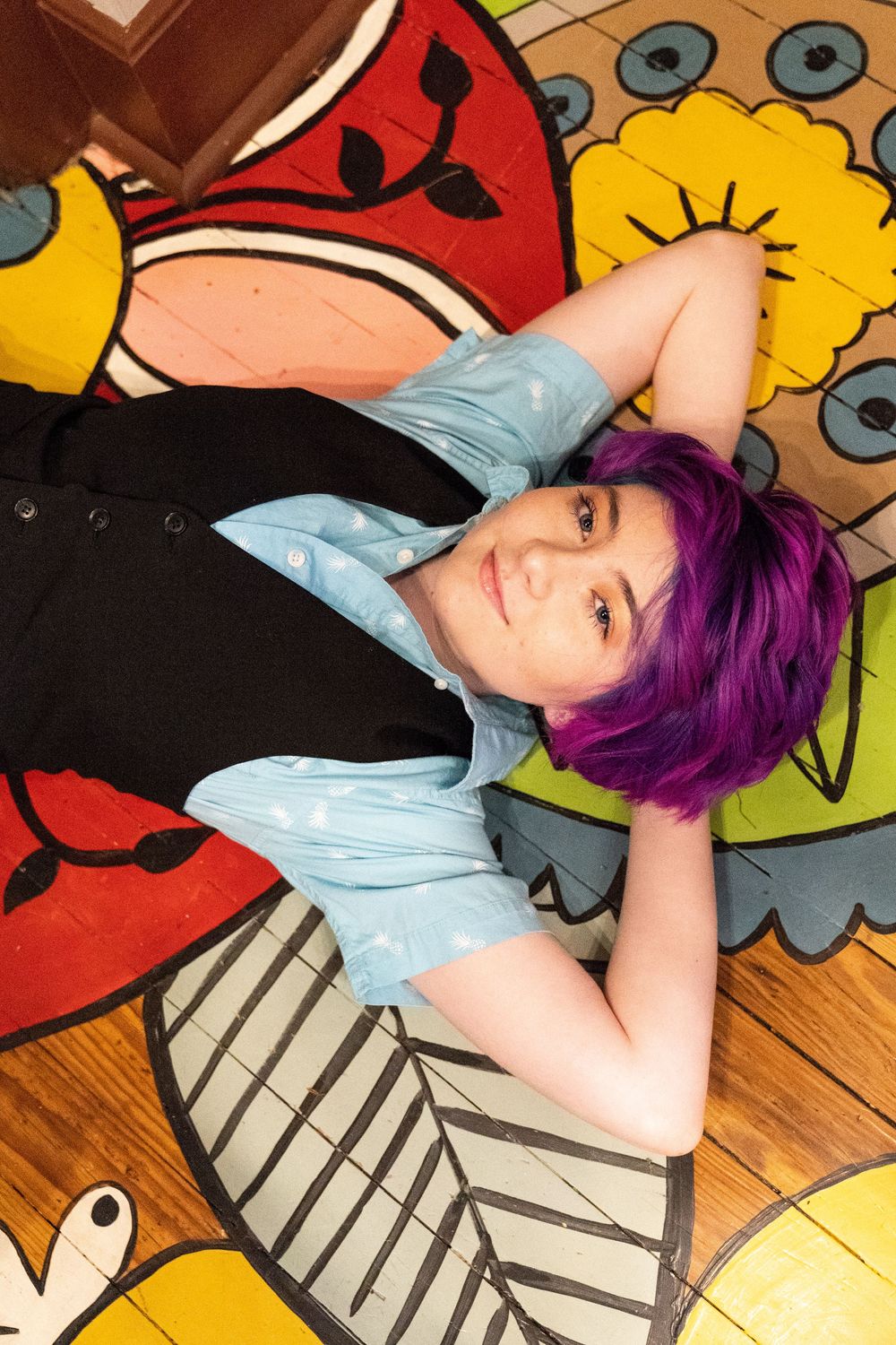 Senior portrait of purple haired girl laying on a colorful floor with her hands behind her head wearing a suit vest.