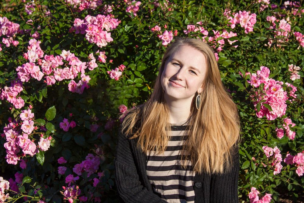 Senior portrait of high school student with long blonde hair, stripped shirt and black sweater in front of a bush of pink flowers.