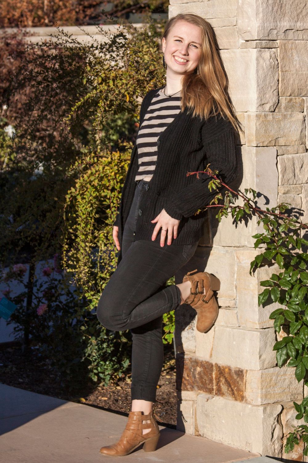 Senior portrait of high school student in black jeans, stripped shirt, and black sweater leaning against rock column.