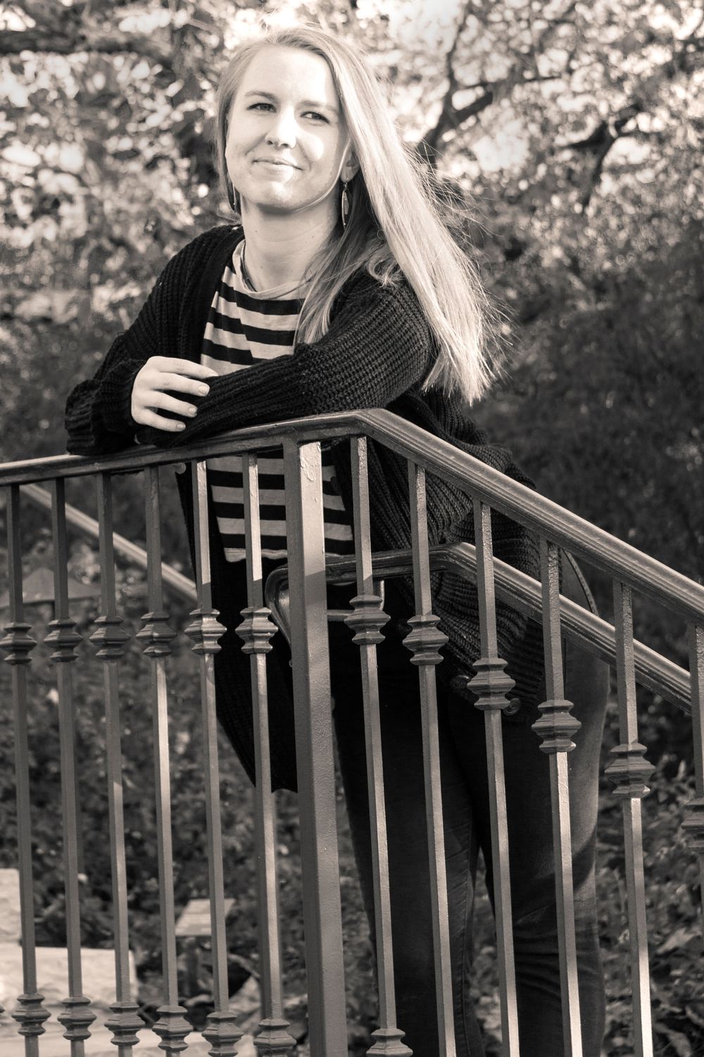 B&W Senior portrait of high school student with long blonde hair in stripped shirt and black sweater leaning on iron railing.