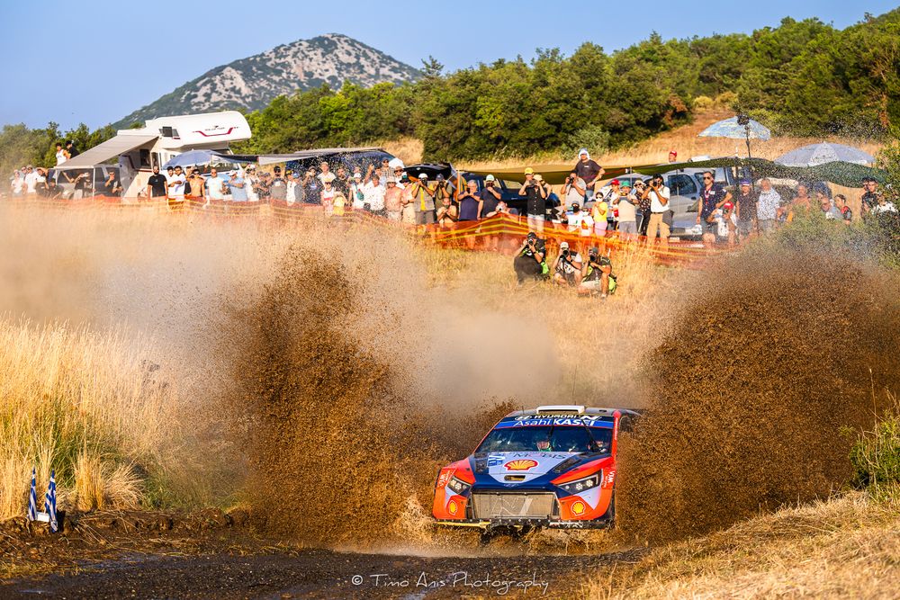 Rally cars kicking up dust clouds while racing through a desert track with spectators watching from the sidelines.