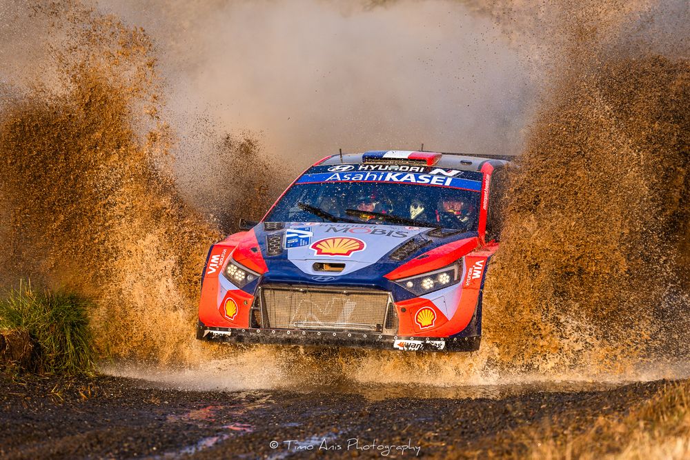 Rally car racing through muddy water in high speed racing action, splashing dirt and debris into the air.