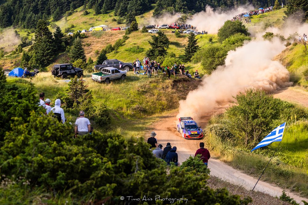 Rally car kicks up dust on mountain course as spectators watch from grassy hillside with Greek flags visible.