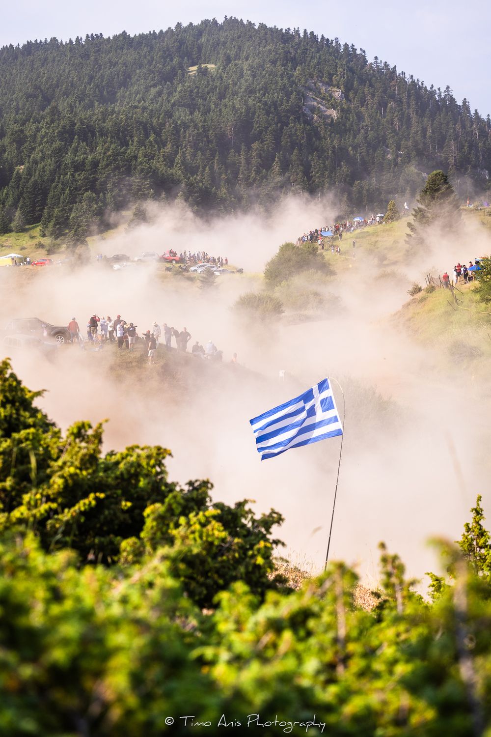 Greek national flag waves in a dusty rally course with mountains and trees visible in the background.