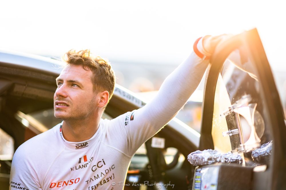 Race car driver in white shirt holds car door open while glowing sunlight creates lens flare effect in background.