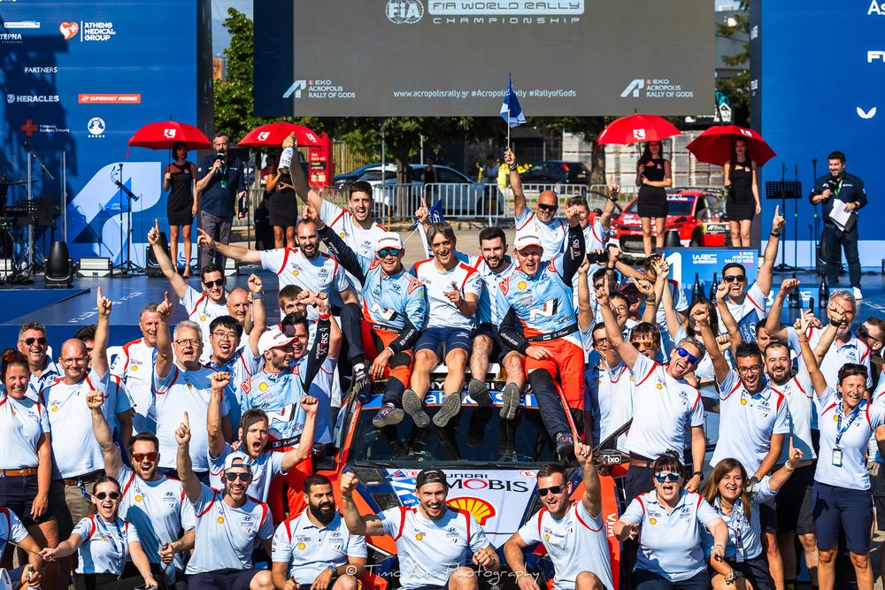 A large group of team members in white uniforms celebrate together on an outdoor podium at a racing event.