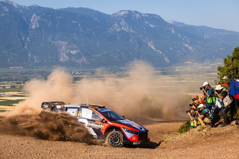 Rally cars racing through mountainous terrain kick up dust clouds as spectators watch from the sidelines during competition.