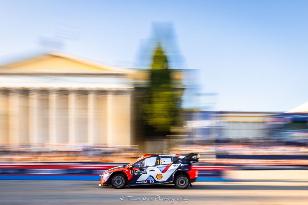 Race car speeds along track creating colorful motion blur effect against bright blue sky and buildings.