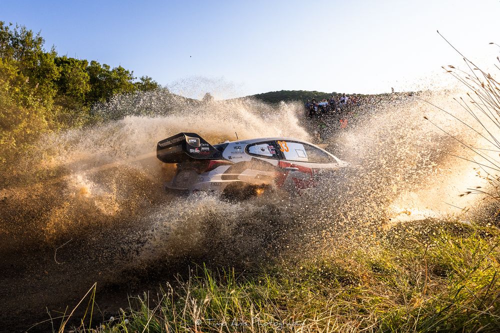 Rally car splashes through deep water and mud at high speed throwing up dramatic spray during off-road race.
