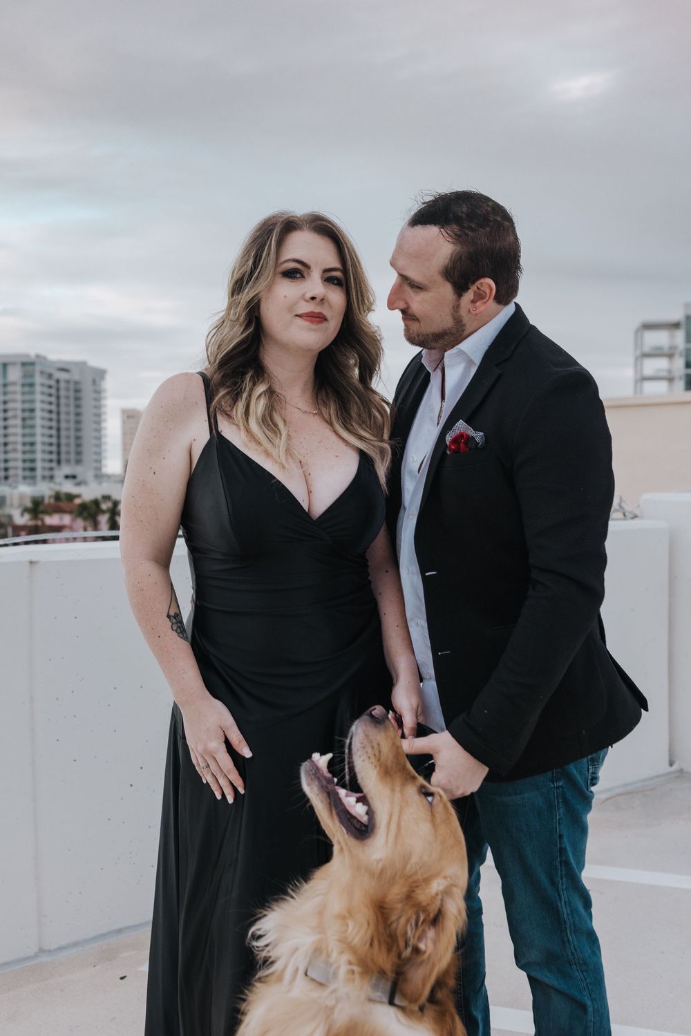 Couple in formal attire with a golden retriever on a rooftop with cityscape in the background.