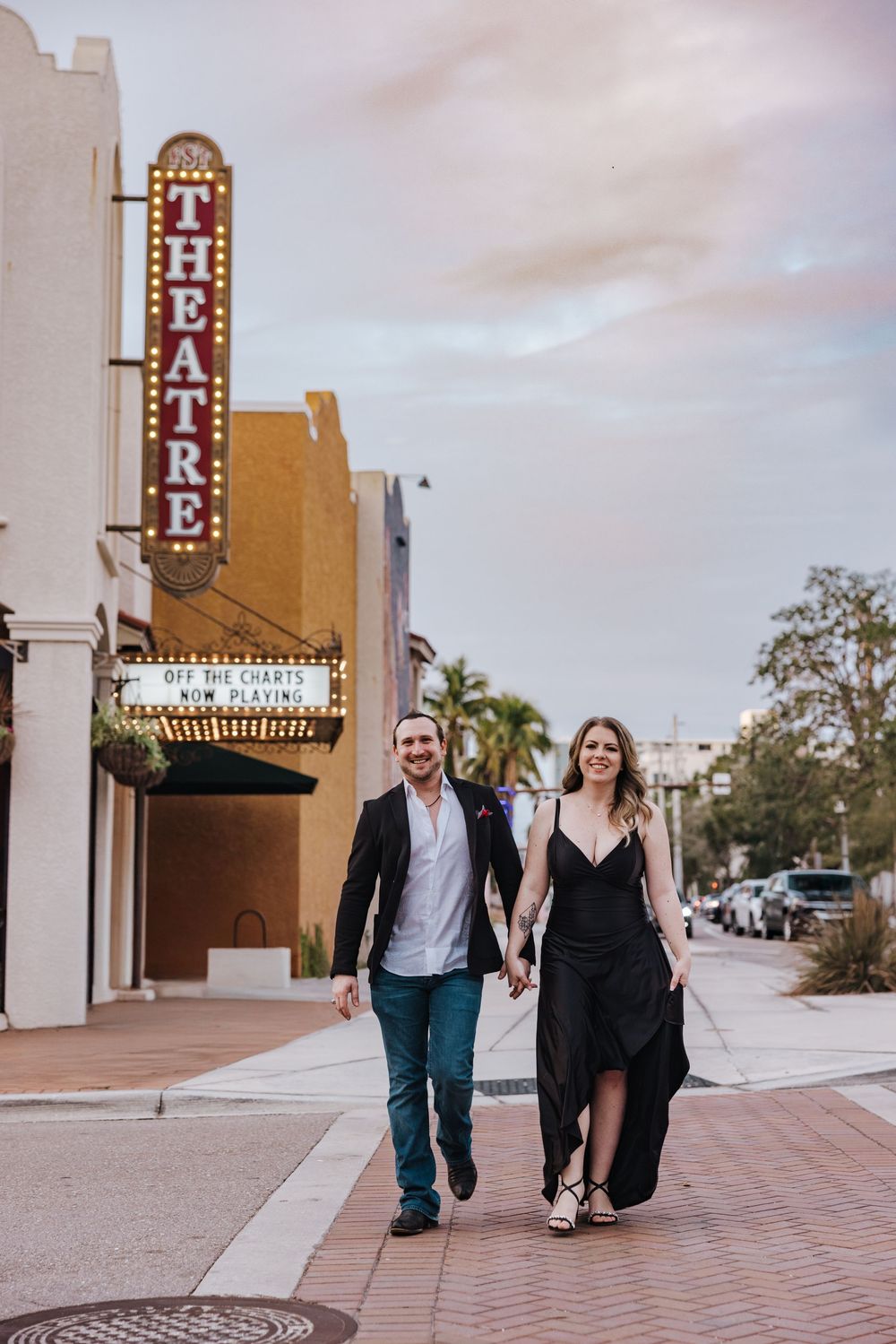 A couple strolls hand in hand on a sidewalk in front of a vintage theatre with a classic marquee sign.