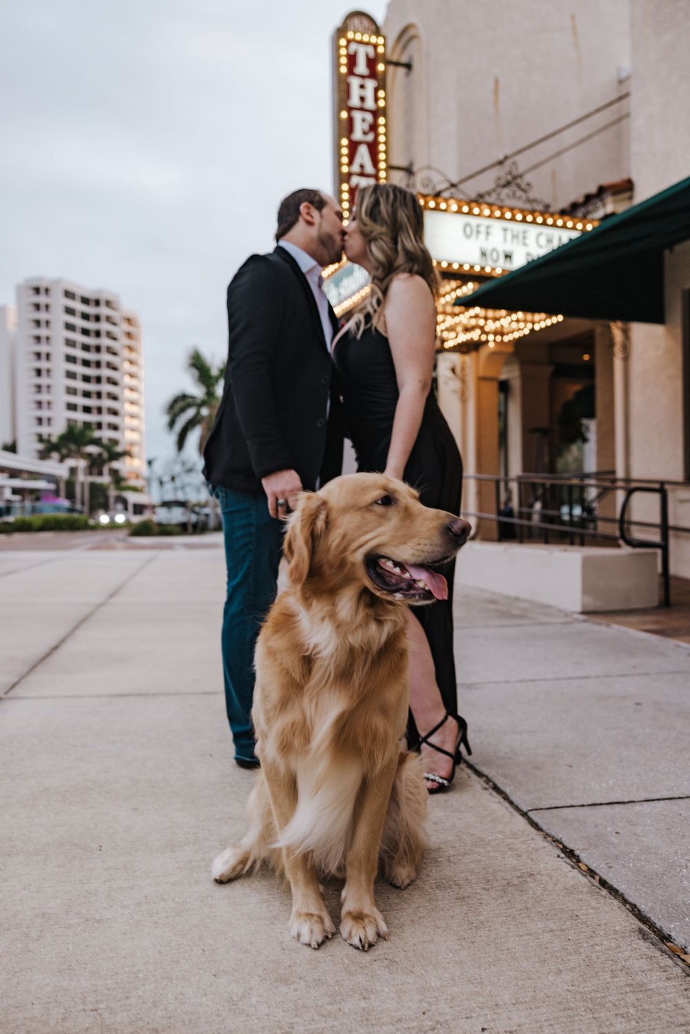 A golden retriever sits obediently outside a building with marquee lights while its owner stands nearby.
