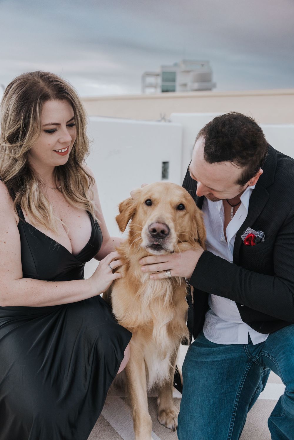 Couple in formal attire pose with their golden retriever on rooftop patio during engagement photo shoot.
