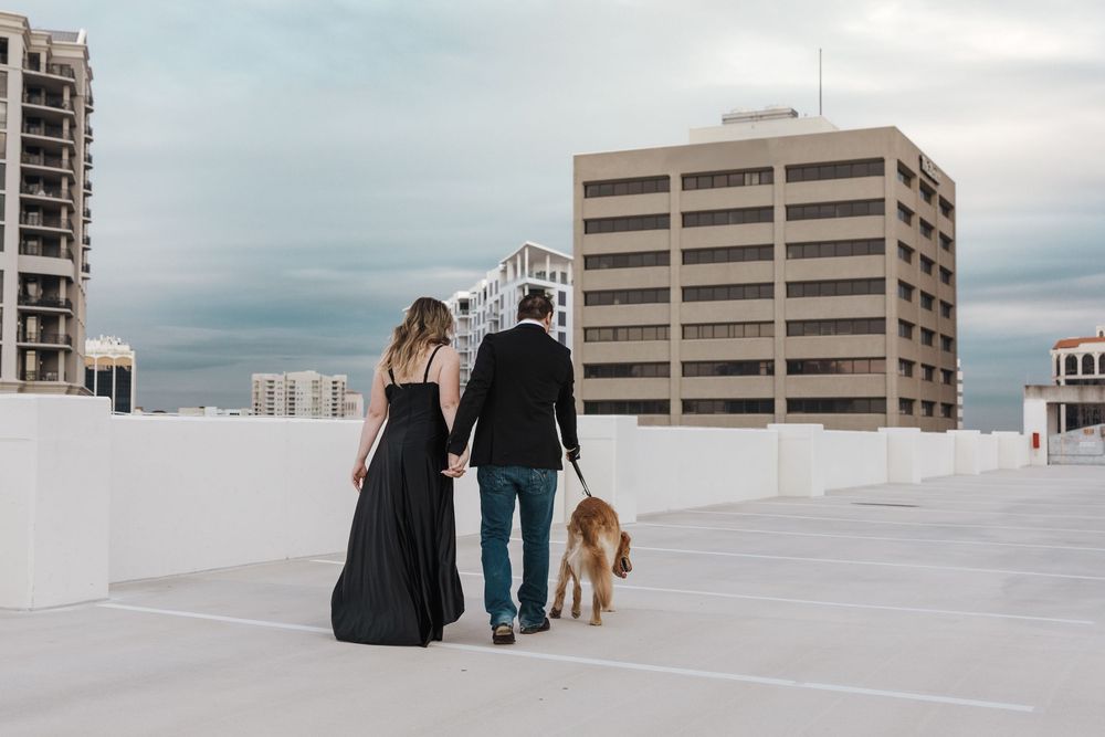 A couple and their dog stand together on a white rooftop overlooking urban buildings on a cloudy day.
