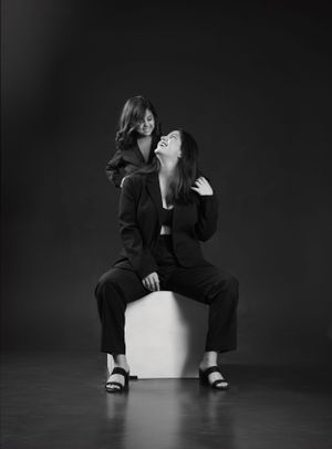 Two figures in black suits sitting on white cube in artistic studio portrait.