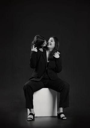 Two people sharing laughter while sitting on white cube against dark background.