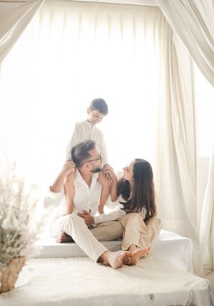 Family enjoying playful interaction on white bedding in a bright room with sheer curtains.