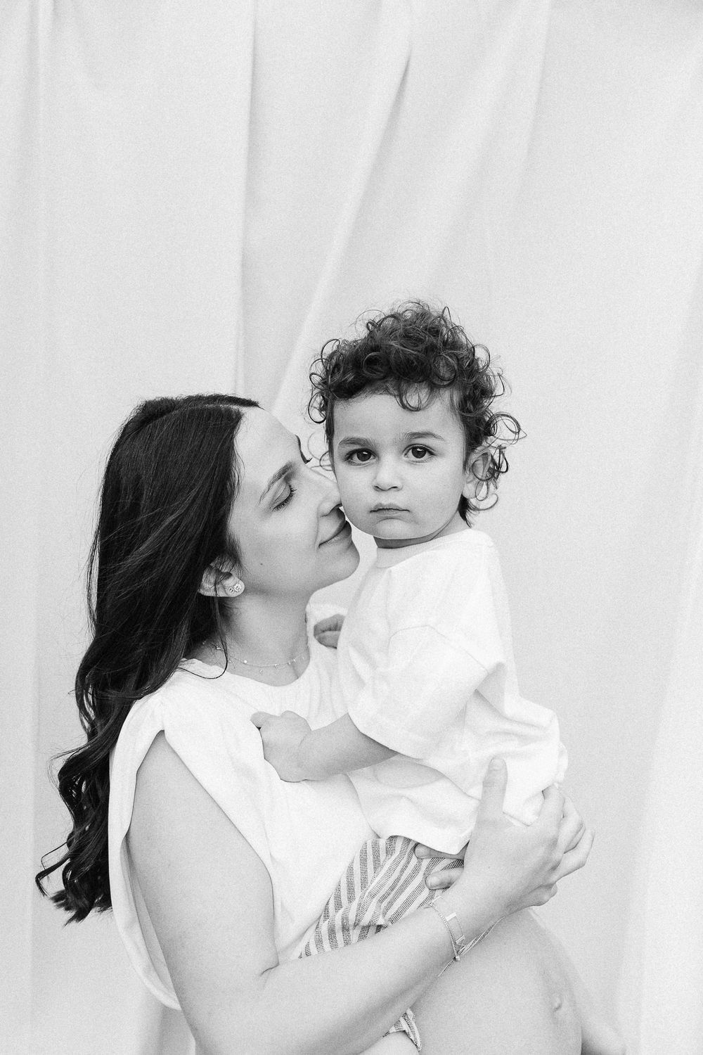 A tender black and white portrait of a mother and young child with curly hair against a light background.