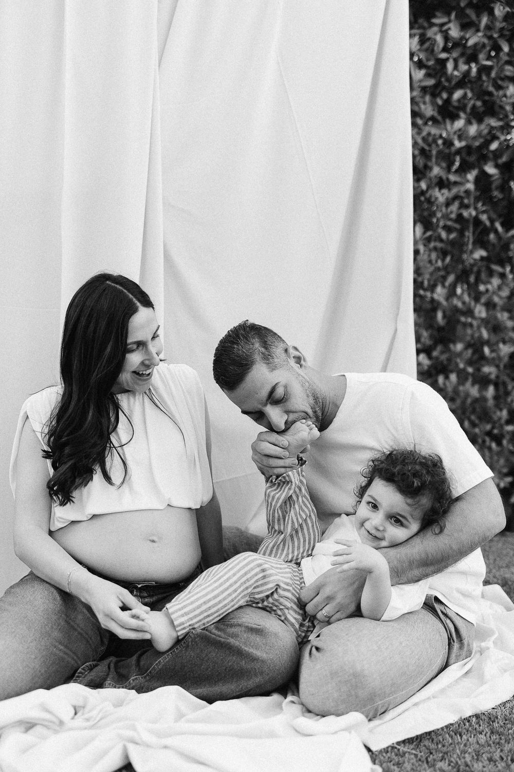 A loving black and white family portrait shows a couple sitting together with their toddler on a white blanket outdoors.
