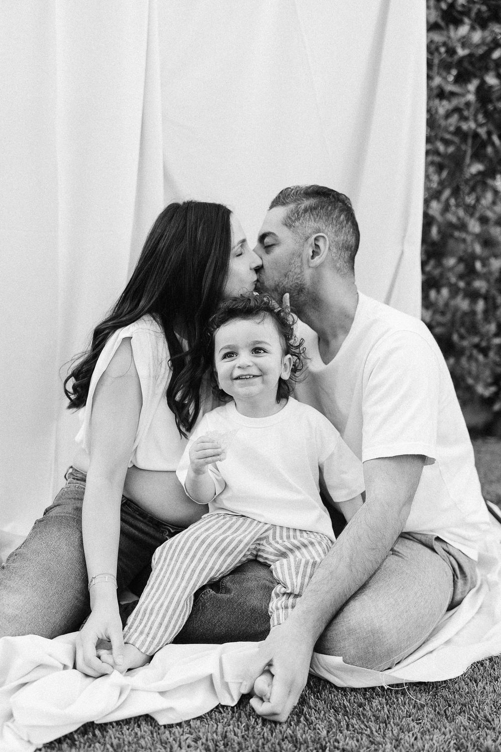 Sweet black and white family portrait showing parents kissing while their child smiles between them.