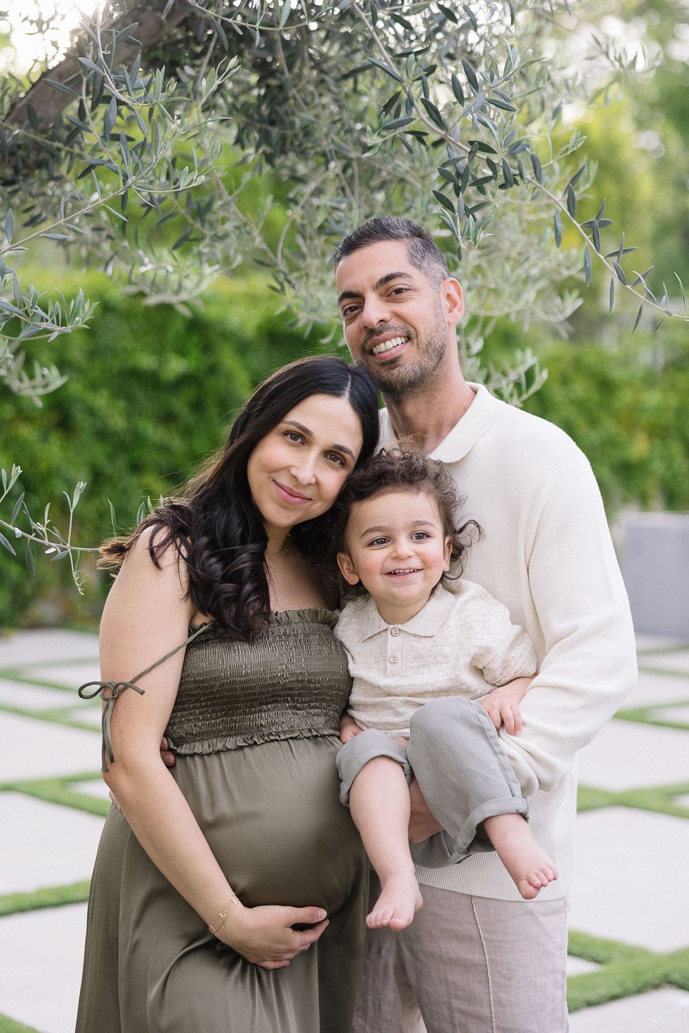 A family poses together under an olive tree with a visible pregnant belly in an olive green dress and casual outdoor setting.