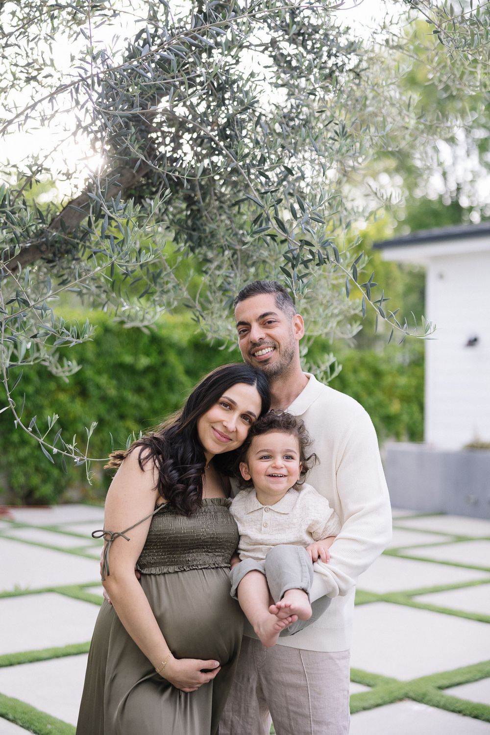 Family poses together in an outdoor garden setting with olive trees and modern landscaping in the background.