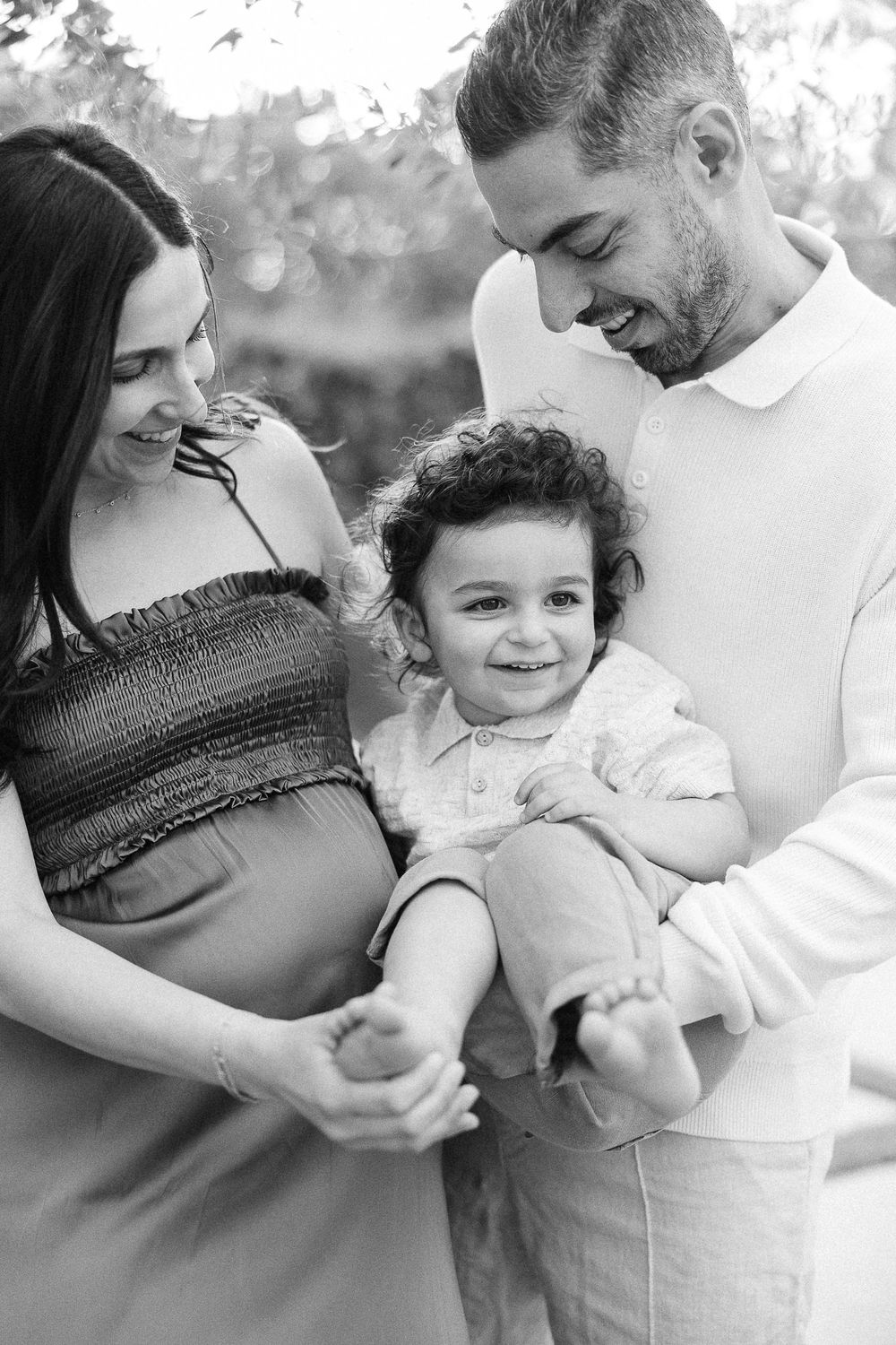 Black and white family portrait series showing intimate moments between parents and baby in white clothing outdoors.