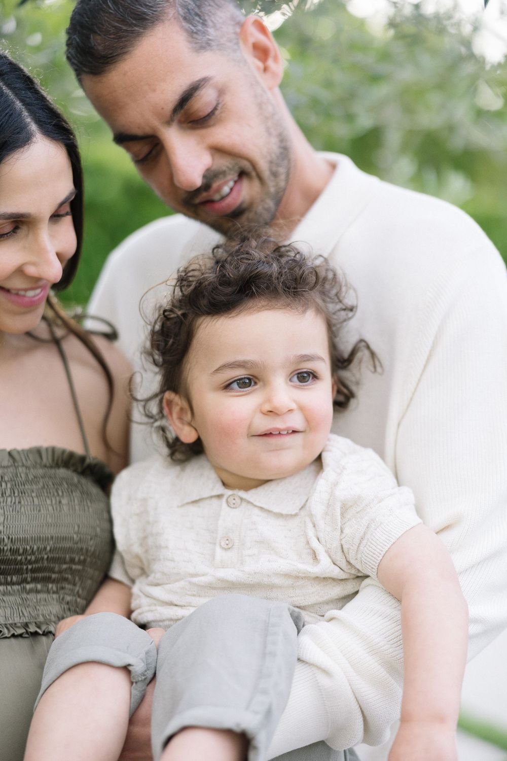 A tender family moment outdoors with a small child with curly hair wearing light colored clothing.