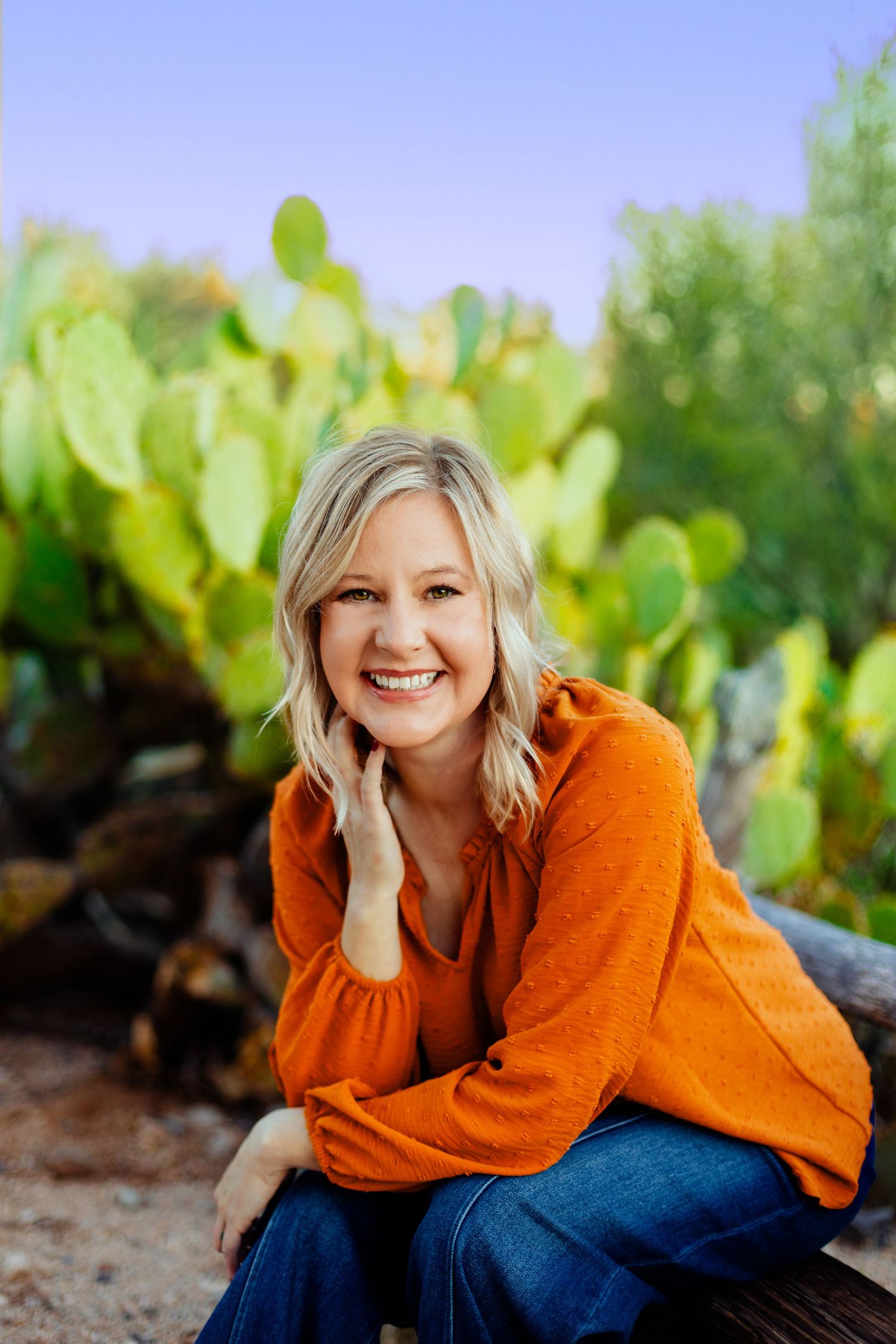 Person in bright orange sweater sits casually against desert landscape with cactus backdrop.