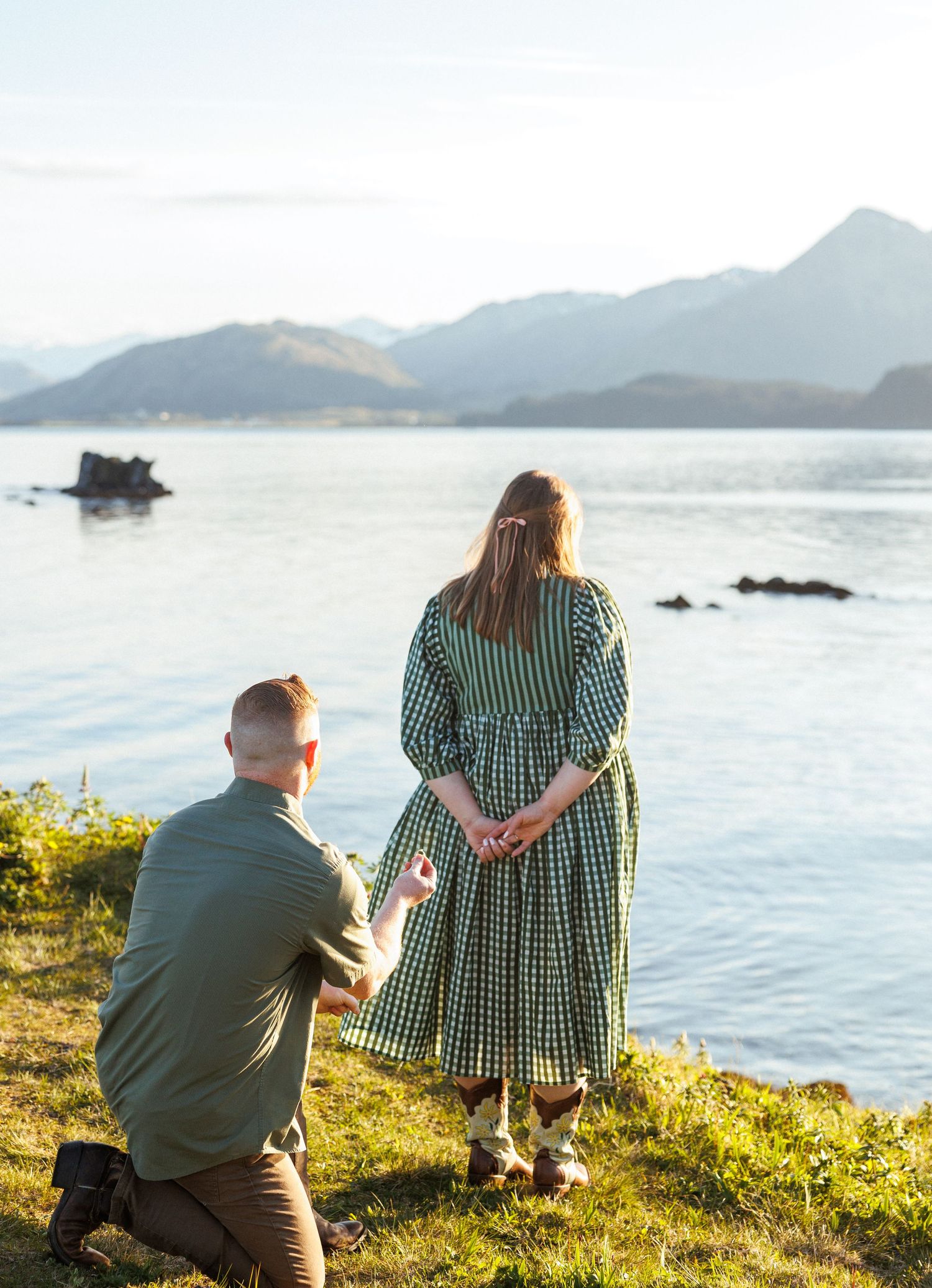 A romantic proposal unfolds along a scenic coastal cliff with mountains and ocean views at golden hour.