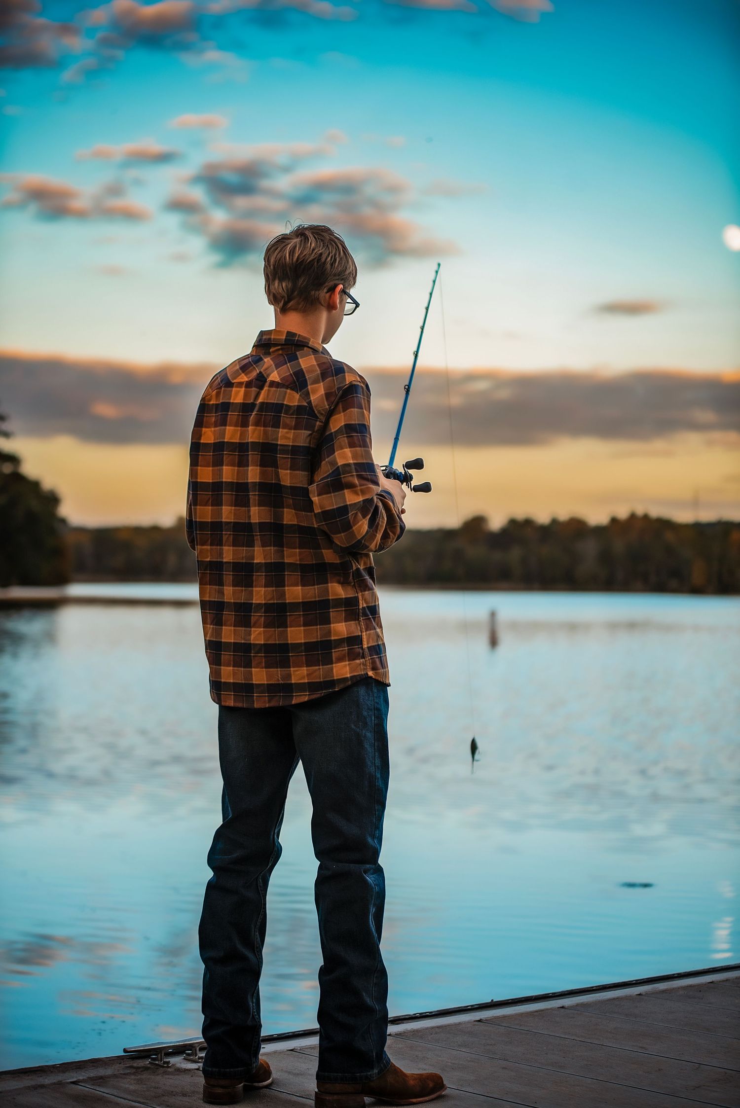 Person in plaid flannel shirt fishing at sunset on a tranquil lake with colorful clouds reflecting on calm waters.