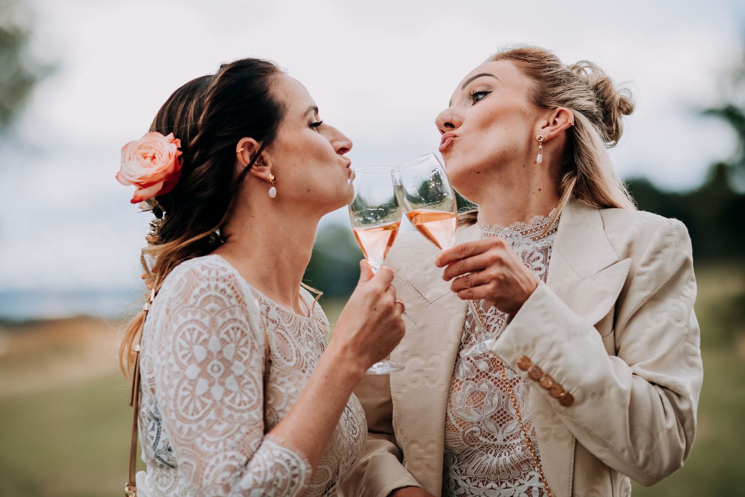 Two brides dressed in lace and cream outfits share playful moment while taking a break from a celebration outside.
