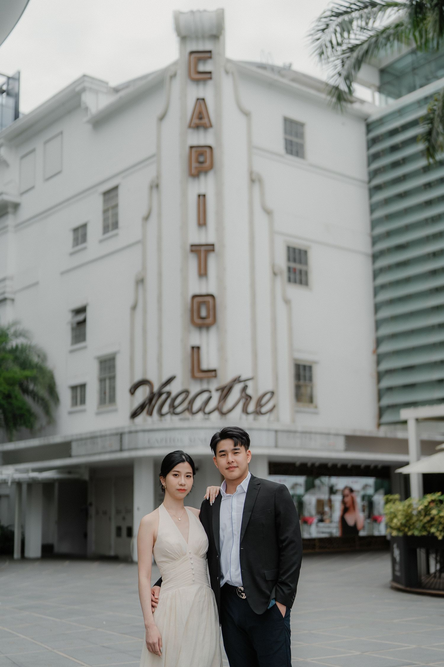 A couple poses in front of the historic white Capitol Theatre building with art deco signage and palm trees.