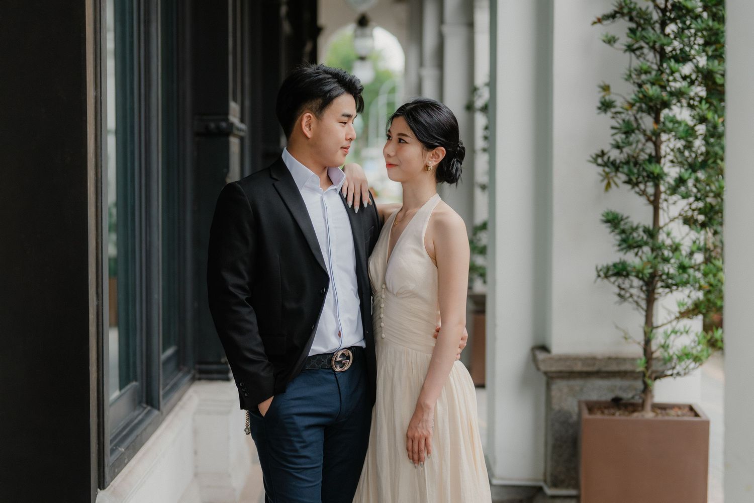 A couple in elegant wedding attire embrace and smile while standing in front of a white building with greenery.