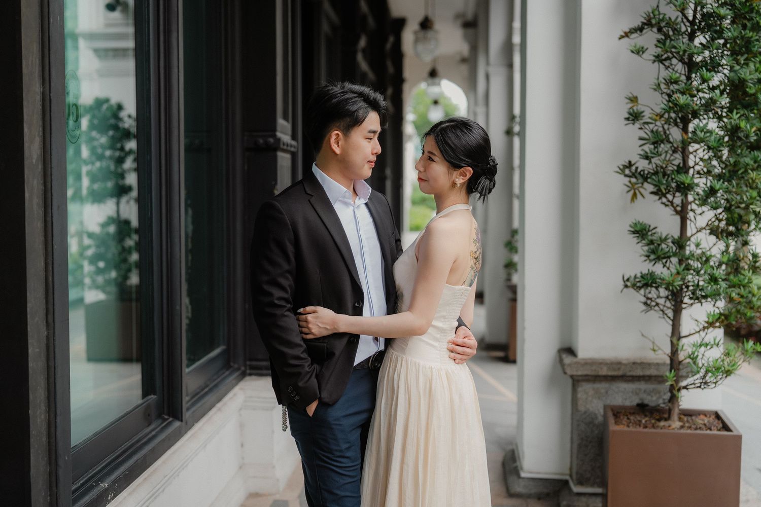 A couple shares a romantic moment in an elegant hallway with potted plants and glass windows.