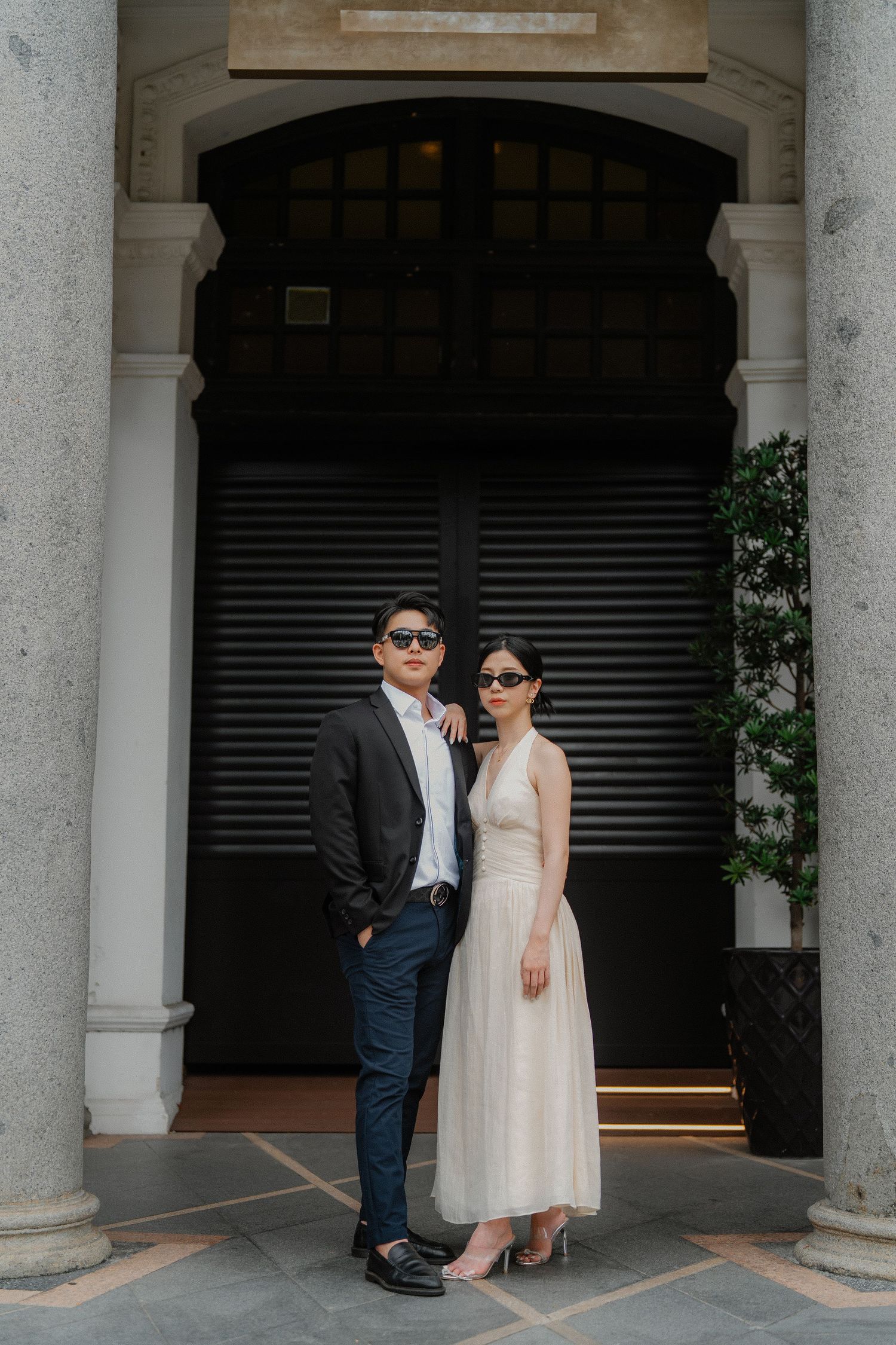 A stylish couple wearing sunglasses poses under an arched doorway in formal wear.