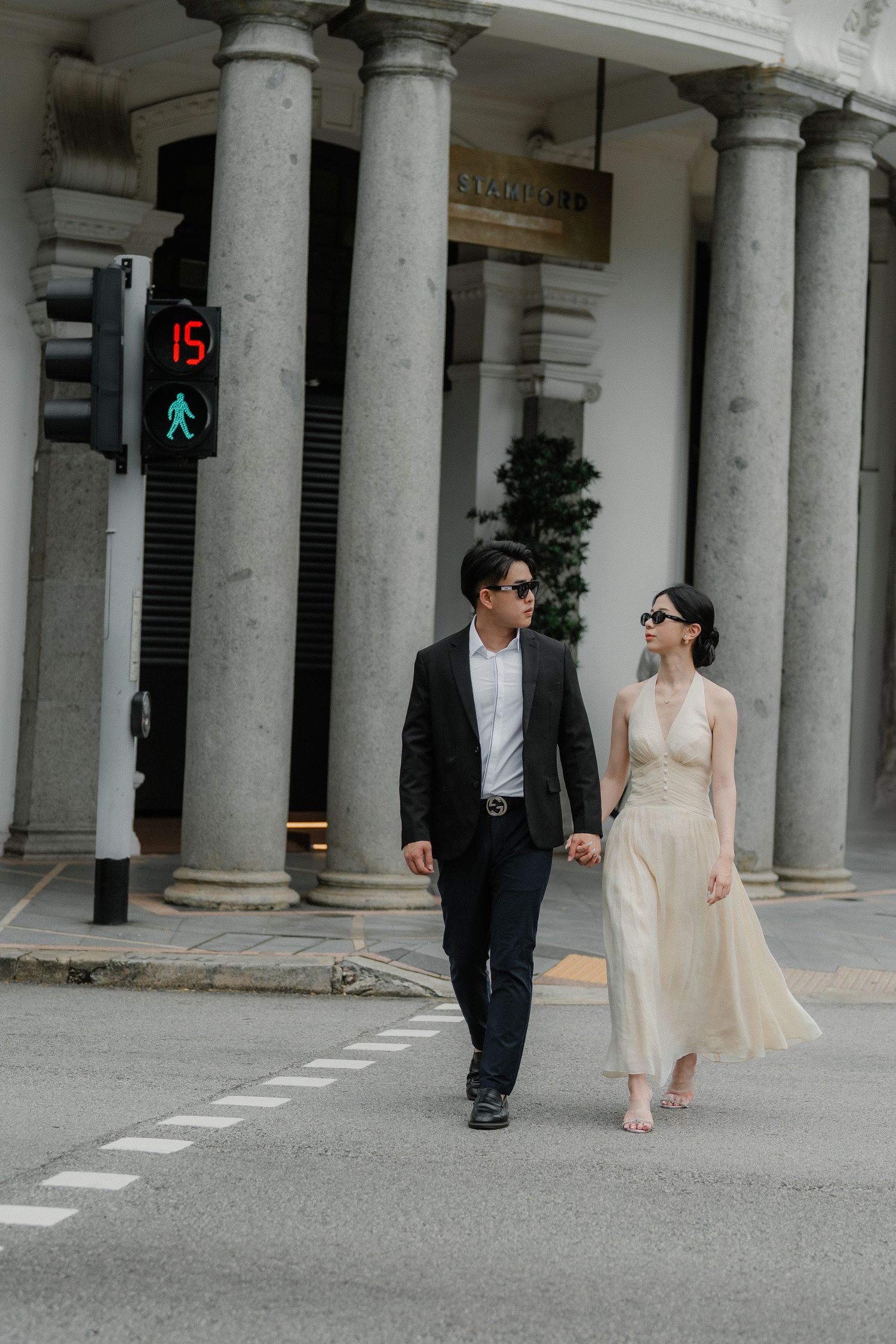 Stylish couple walking hand in hand past classic colonial architecture with traffic light in background.