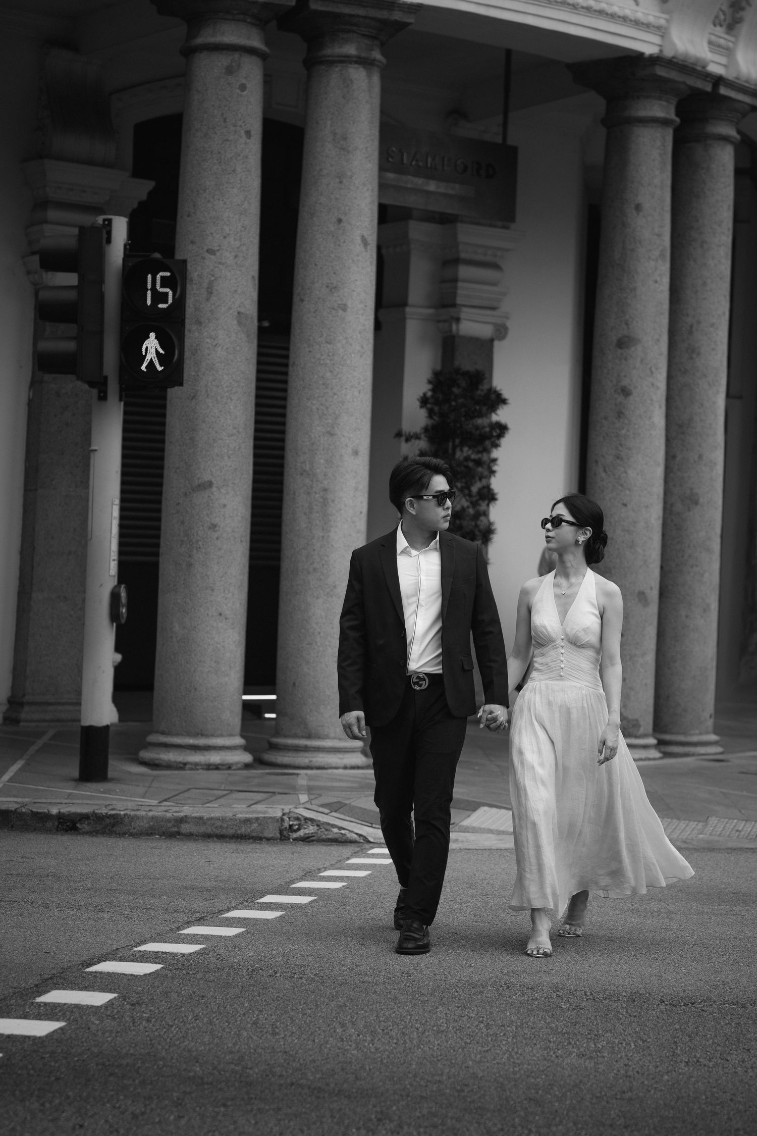 A couple in formal attire walks hand in hand along a colonnade in black and white photography.