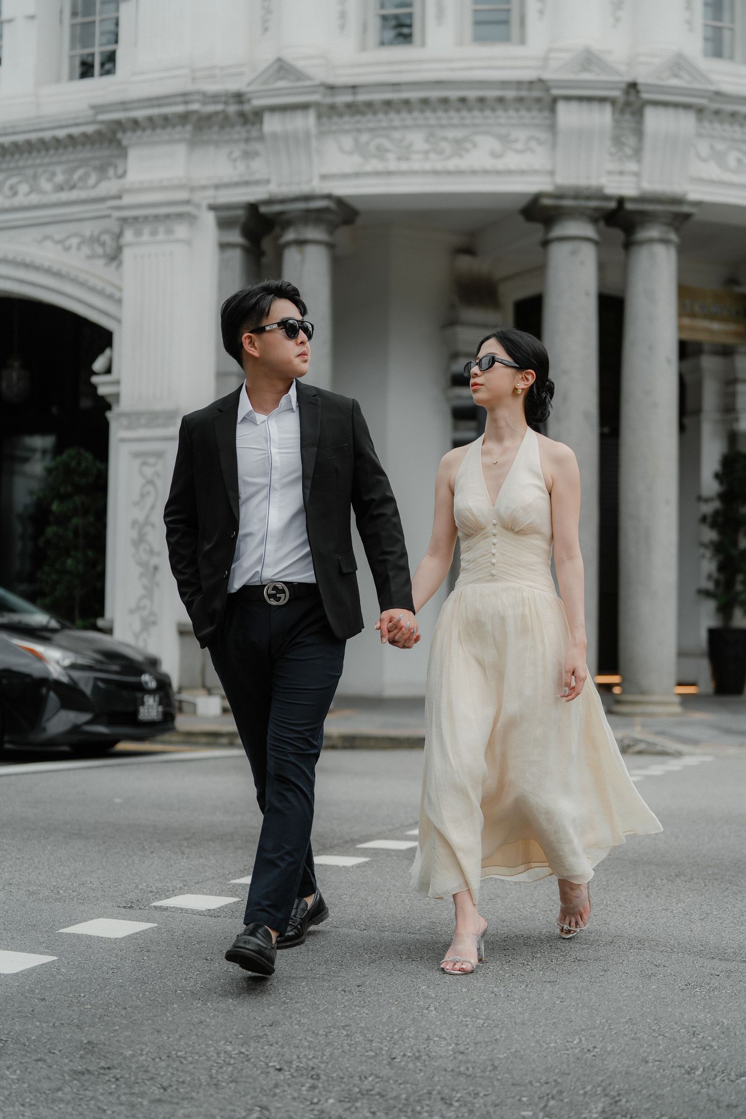 A couple holds hands while walking along a street by a classic white colonial building with pillars.