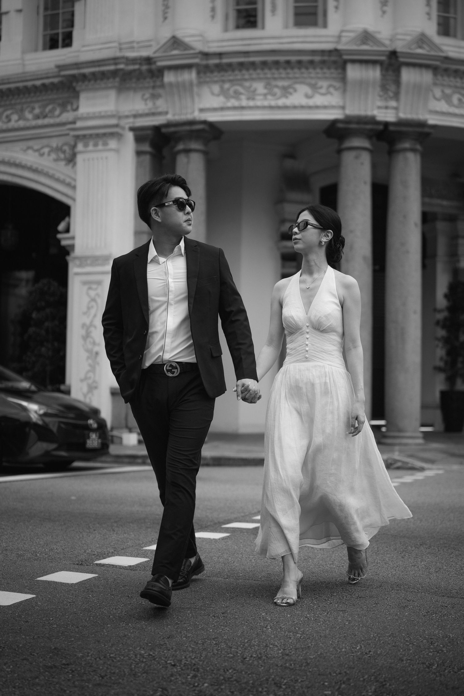 A stylish couple in formal wear walks hand in hand on a street with classic architecture.