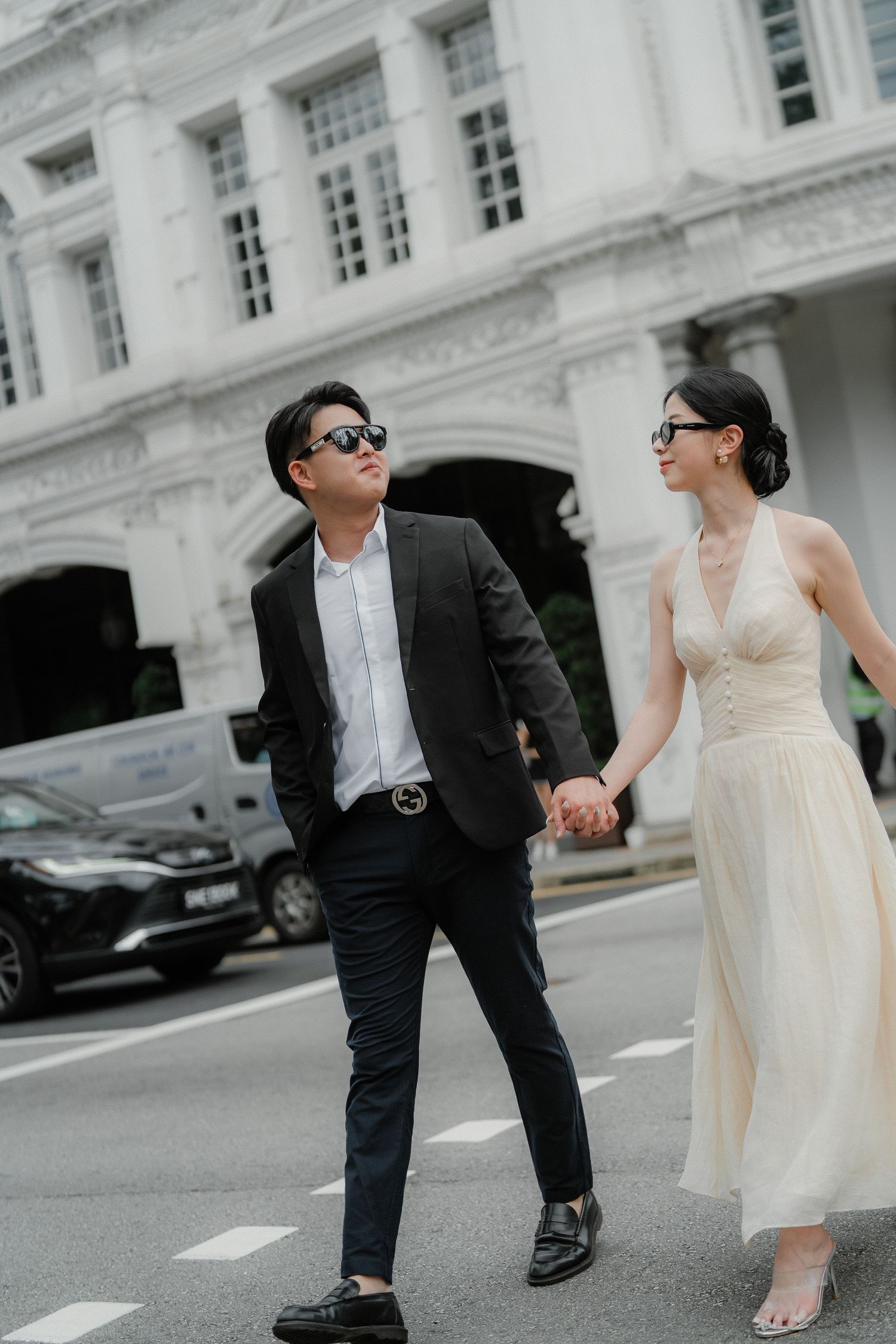 A couple in formal attire strolls hand in hand outside an elegant white colonial building.
